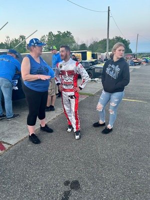 Three people, including a race car driver in suit, stand outdoors.